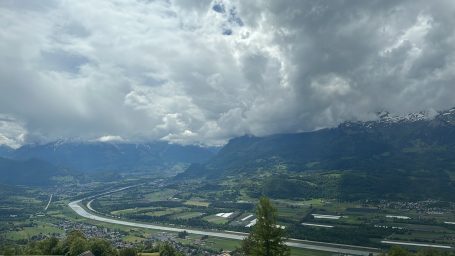 Berglandschaft mit Wolken und Fluss im Tal, umgeben von grünen Wiesen.